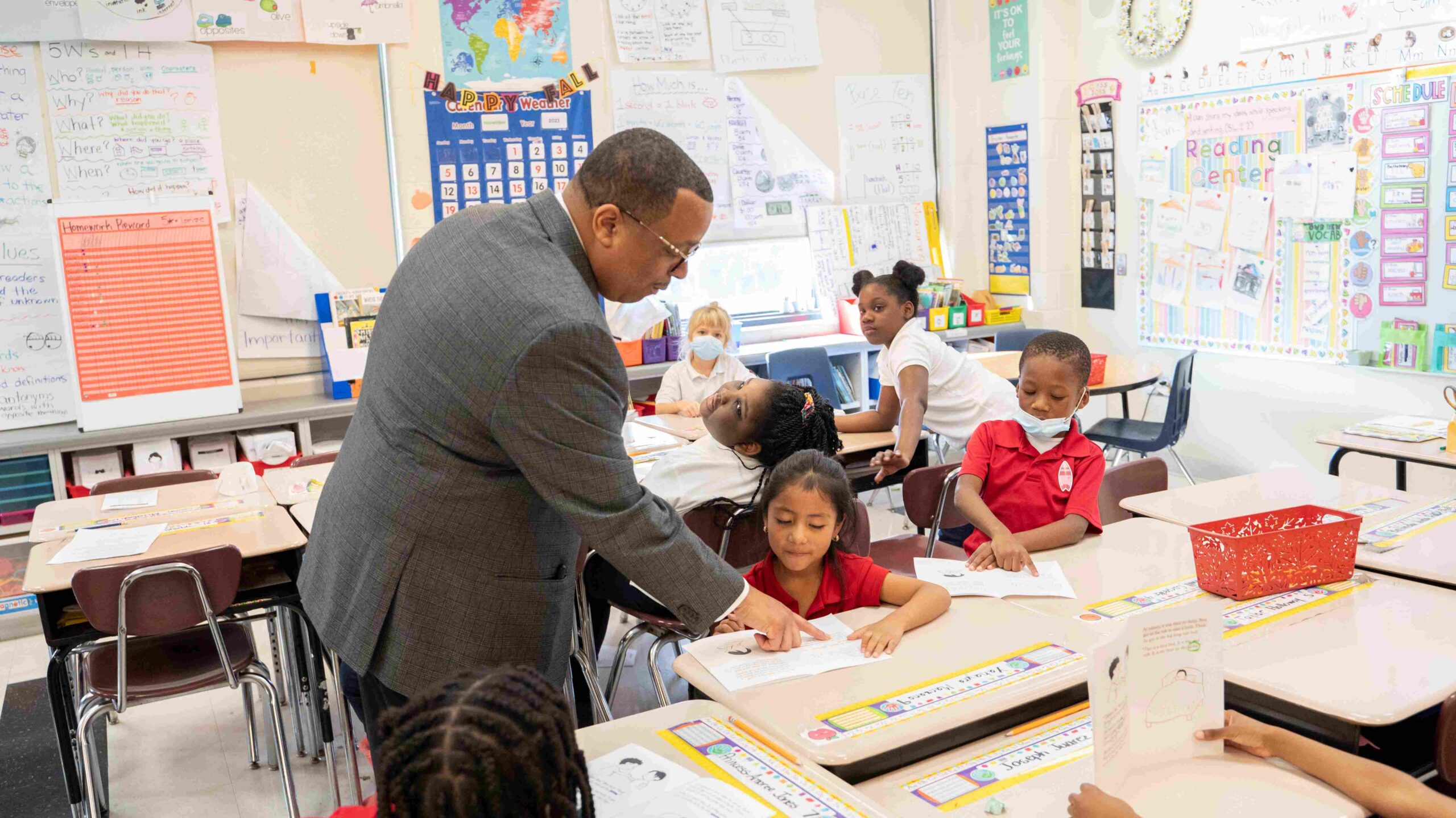 A principal in a gray suit points to a piece of paper that a student is reading in an elementary school classroom. The student sits at a cluster of four desks with other students.