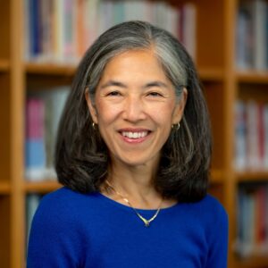 A woman in a royal blue blouse smiles for the camera. She has graying, shoulder-length, black hair. She is wearing small golden circular earrings and a simple golden necklace. She is in front of bookshelves.